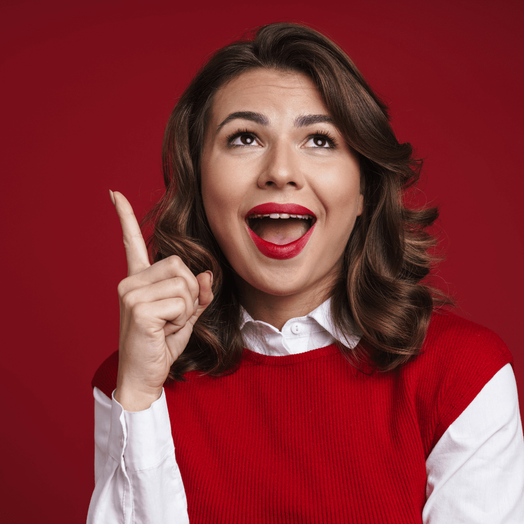 Picture of a smiling, excited woman, wearing a red vest, looking and pointing up in front of a red background.